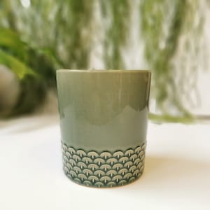 May include: A sage green ceramic cup with a textured, patterned base. The cup has a smooth, glossy finish and a cylindrical shape. The base features a repeating, scalloped design. The cup is likely used for beverages or as a decorative item.