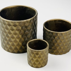 May include: Three bronze-coloured cylindrical planters with a textured, geometric pattern. The planters vary in size, with the largest measuring approximately 15 cm in height and 13 cm in diameter. The smallest planter is about 8 cm tall and 8 cm in diameter.