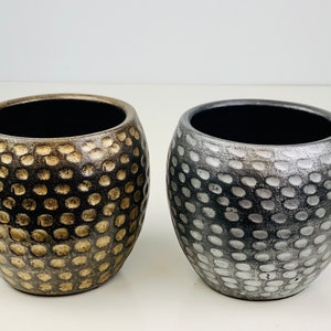 May include: Two round ceramic planters with a textured, dimpled surface. One planter is gold and the other is silver.