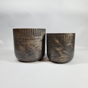 May include: Two round, dark brown metal planters with a textured finish. The larger planter is slightly taller than the smaller planter.