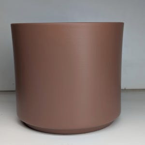 May include: A brown cylindrical plant pot with a rounded base. The pot has a smooth, matte finish, suitable for indoor or outdoor use. The neutral colour complements various decor styles. The pot is approximately 15 cm in diameter.