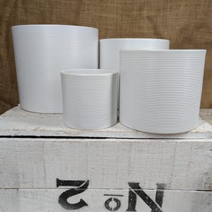 May include: Four white ceramic plant pots of different sizes, each featuring a horizontal ribbed design. They are displayed on a white wooden crate with the number "2" and the letter "N" stencilled in black.