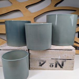 May include: Four teal-coloured, ribbed ceramic plant pots of different sizes. They are arranged on a white wooden crate. The background features a decorative wooden panel with cut-out shapes. These pots are suitable for indoor or outdoor use. Measurements are in centimetres.
