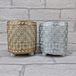 May include: Two decorative concrete planters with a geometric pattern. The planter on the left is gold and the planter on the right is silver.