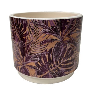 May include: A ceramic planter with a purple and brown tropical leaf pattern. The planter has a white rim and a white base.