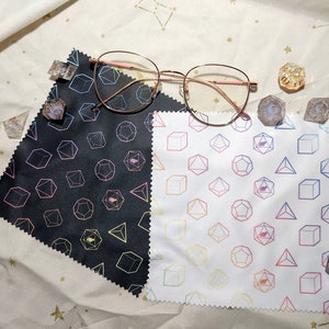May include: Two microfiber cleaning cloths with a geometric pattern of dice. One cloth is black with a rainbow pattern and the other is white with a rainbow pattern. A pair of gold-rimmed glasses and several dice are scattered around the cloths.