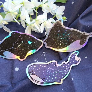 May include: Three iridescent holographic stickers featuring a whale, a manta ray, and a shark, each with a constellation design. The stickers are on a blue fabric background.