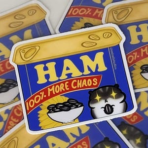 May include: A blue and yellow sticker with the text "HAM 100% MORE CHAOS" and a cartoon hamster with big eyes. The sticker is designed to look like a can of ham.