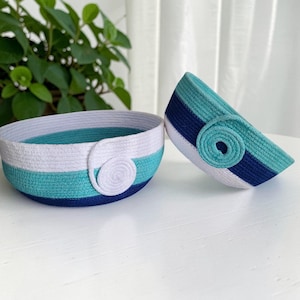 May include: Two round baskets crafted from coiled rope in horizontal stripes of white, turquoise, and navy blue. Each basket features a decorative circular rope accent on the side, with one basket positioned upright and the other tilted.