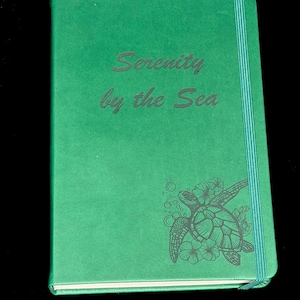 May include: A green journal with the words "Serenity by the Sea" embossed on the cover. A turtle and floral design is on the lower right corner. The journal has a teal elastic band.