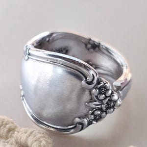 May include: A silver-toned metal ring crafted from a repurposed spoon, featuring a smooth, rounded front surface and intricate embossed floral detailing along the sides and band, showcasing a polished metallic finish throughout the piece.