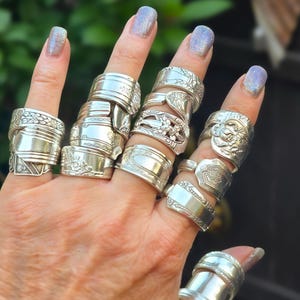 May include: A collection of silver-toned rings crafted from repurposed vintage silverware handles. Each ring features unique embossed floral, scrollwork, or geometric patterns, with varying widths and designs worn across multiple fingers.