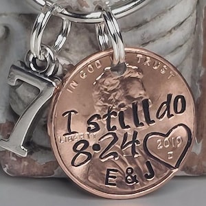 May include: A silver-toned metal keychain featuring a circular copper-colored coin charm and a silver number seven charm. The coin is stamped with the text I still do, the date 8-24, the year 2019, the initials E & J, and a heart shape.