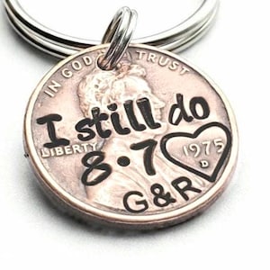 May include: A copper-colored metal keychain featuring a modified United States penny. The coin is engraved with the black text I still do, the date 8.7, the initials G&R, and a heart shape containing the year 1975 and the letter D.