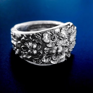 May include: A silver-toned spoon ring featuring an intricate embossed floral pattern across the entire outer surface. The design displays detailed petals, leaves, and stems in a relief style, wrapping around the band to form a continuous metallic floral motif.