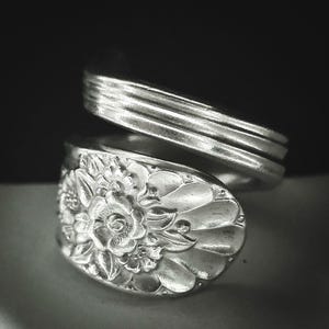 May include: A silver-toned metal ring crafted from a repurposed spoon. The band features a smooth, multi-ridged design that wraps around the finger, while the focal point is the bowl of the spoon, embossed with a detailed floral pattern.