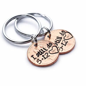 May include: Two silver-toned metal key rings, each attached to a copper-colored penny. The pennies are stamped with the black text "I still do" followed by "5.12" and a heart shape containing the year "2017" engraved on the surface.