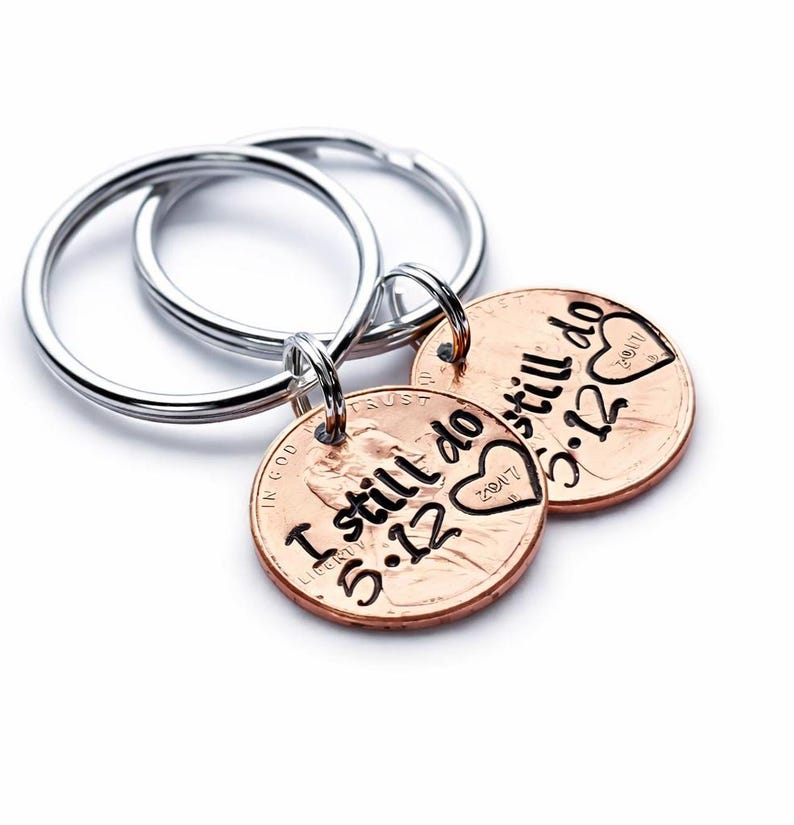 May include: Two silver-toned metal key rings, each attached to a copper-colored penny. The pennies are stamped with the black text "I still do" followed by "5.12" and a heart shape containing the year "2017" engraved on the surface.