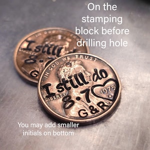 May include: Two copper-colored United States one-cent coins featuring stamped black text. The coins are engraved with the words I still do, the date 8.7, the year 1975 inside a heart shape, and the initials G&R, with Liberty and In God We Trust.