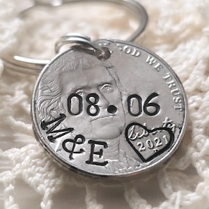 May include: A silver-toned metal keychain featuring a modified United States nickel coin. The coin is stamped with the date 08.06, the letters M&E, and a heart shape containing the year 2021. The text GOD WE TRUST is visible on the coin.