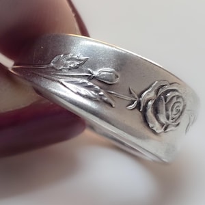 May include: A silver-toned metal ring featuring an embossed floral design that includes a blooming rose, a small bud, and slender leaves. The band has a smooth, polished finish with the intricate botanical pattern raised along the outer surface.