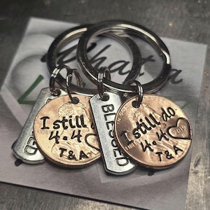 May include: Two silver-toned metal keychains featuring circular copper-colored coins stamped with the text I still do, 4.4, T&A, and a heart symbol with 2019. Each keychain includes a rectangular silver-toned metal tag stamped with the word BLESSED.