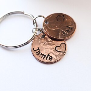 May include: A keychain with two copper pennies. One penny has the word "Jamie" engraved on it and a heart with the year 2025. The other penny has the word "Dad" engraved on it and a star.