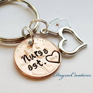 May include: A copper penny keychain with the words "Nurse est." and the year 2019 engraved on it. A silver heart charm and a small silver charm of a nurse's hat are attached to the keychain.