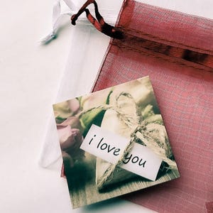 May include: A small, rustic, heart-shaped burlap gift bag with a handwritten tag that says "i love you". The bag is surrounded by a floral arrangement with a pink rose.