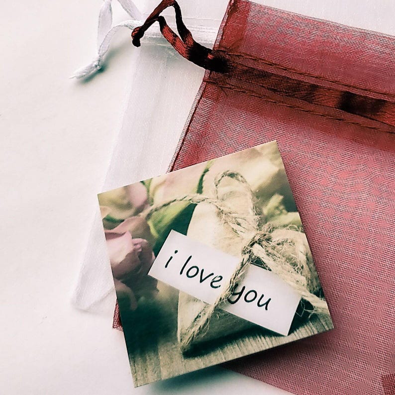 May include: A small, rustic, heart-shaped burlap gift bag with a handwritten tag that says "i love you". The bag is surrounded by a floral arrangement with a pink rose.