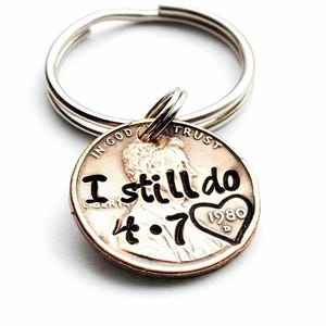 May include: A silver-toned metal keyring attached to a copper-colored United States penny. The coin is stamped with the black text "I still do", the date "4.7", and a heart shape containing the year "1980" over the profile of Abraham Lincoln.