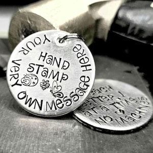 May include: Close-up of two silver metal round tags. The larger tag has the words "YOUR VERY OWN MESSAGE HERE" and "HAND STAMP" with illustrations of a ladybug, butterfly, and flower. The smaller tag reads "Each letter 1 at a time, NO MACHINE".