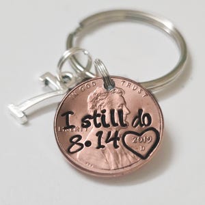 May include: A metal keychain featuring a copper-colored penny stamped with the text I still do, the date 8.14, and a heart shape containing the year 2019. A silver-toned number 7 charm is attached to the split ring alongside the penny.
