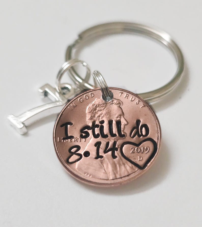 May include: A metal keychain featuring a copper-colored penny stamped with the text I still do, the date 8.14, and a heart shape containing the year 2019. A silver-toned number 7 charm is attached to the split ring alongside the penny.
