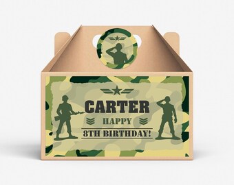 以下が含まれることがあります： 迷彩柄の茶色の段ボール箱に「Carter Happy 8th Birthday!」の文字が書かれています。箱の上部に切り抜かれた取っ手があり、迷彩柄の丸いステッカーには兵士のシルエットが描かれています。