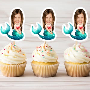 May include: Three cupcakes decorated with colorful sprinkles and topped with mermaid-themed cake toppers. Each topper features a mermaid with a turquoise tail and a girl's face.