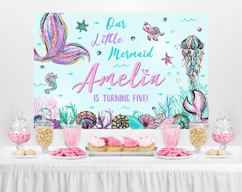 May include: A blue and green mermaid-themed party backdrop with the text "Our Little Mermaid Amelia is turning five!"  The backdrop features a mermaid tail, a starfish, a seahorse, and a jellyfish.