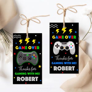 K&ouml;nnte beinhalten: Zwei schwarz-wei&szlig;e Geschenkanh&auml;nger mit einem Gamecontroller-Motiv und dem Text "GAME OVER" in bunten Buchstaben. Der Text "Danke f&uuml;rs Zocken mit mir!" befindet sich unter dem Controller-Motiv. Der Name "ROBERT" befindet sich unter dem Text.