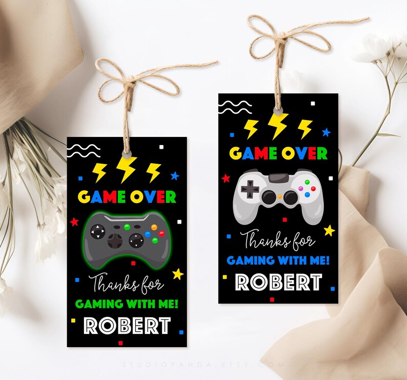 K&ouml;nnte beinhalten: Zwei schwarz-wei&szlig;e Geschenkanh&auml;nger mit einem Gamecontroller-Motiv und dem Text "GAME OVER" in bunten Buchstaben. Der Text "Danke f&uuml;rs Zocken mit mir!" befindet sich unter dem Controller-Motiv. Der Name "ROBERT" befindet sich unter dem Text.