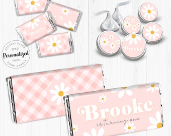 Op de afbeelding: Printbare etiketten voor chocolade reepjes, met een roze en wit ruitjespatroon en madeliefjes. De etiketten bevatten de naam "Brooke" en de tekst "wordt één jaar".