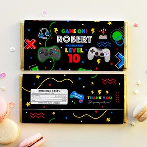 K&ouml;nnte beinhalten: Ein schwarzer und bunter Schokoladenriegel-Wrapper mit Videospiel-Thema. Der Wrapper zeigt einen Gamecontroller, einen Joystick und den Text "GAME ON! ROBERT HAS REACHED LEVEL 10". Die R&uuml;ckseite des Wrappers hat ein N&auml;hrwert-Label und den Text "THANK YOU for gaming with us!"