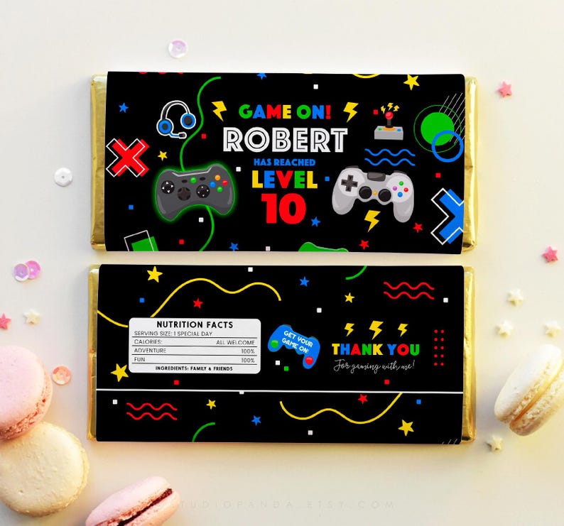 K&ouml;nnte beinhalten: Ein schwarzer und bunter Schokoladenriegel-Wrapper mit Videospiel-Thema. Der Wrapper zeigt einen Gamecontroller, einen Joystick und den Text "GAME ON! ROBERT HAS REACHED LEVEL 10". Die R&uuml;ckseite des Wrappers hat ein N&auml;hrwert-Label und den Text "THANK YOU for gaming with us!"