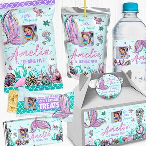 May include: A set of party favor bags, a juice box, a water bottle, and a favor box decorated with a mermaid theme. The bags and boxes feature a mermaid tail with glitter, seashells, and the text "Our Little Mermaid Amelia is turning five!" The water bottle has a similar design with the text "Amelia is turning five!" The juice box has a mermaid tail with glitter and the text "Our Little Mermaid Amelia is turning five!"