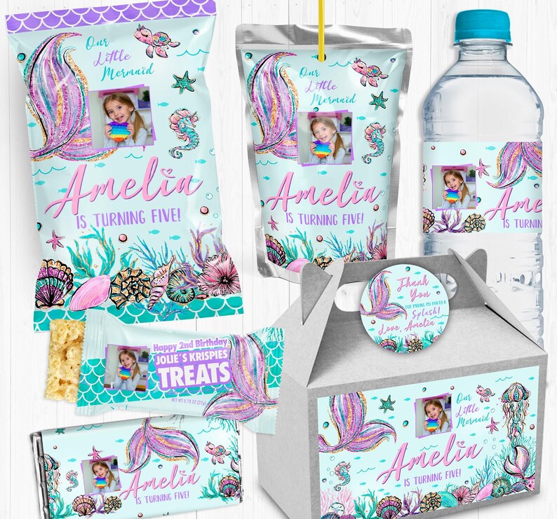 May include: A set of party favor bags, a juice box, a water bottle, and a favor box decorated with a mermaid theme. The bags and boxes feature a mermaid tail with glitter, seashells, and the text "Our Little Mermaid Amelia is turning five!" The water bottle has a similar design with the text "Amelia is turning five!" The juice box has a mermaid tail with glitter and the text "Our Little Mermaid Amelia is turning five!"