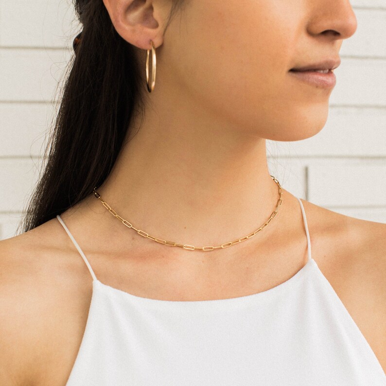 May include: A gold-colored paperclip chain necklace and matching hoop earrings. The jewelry is worn on a person wearing a white spaghetti strap top. The background is a white brick wall.