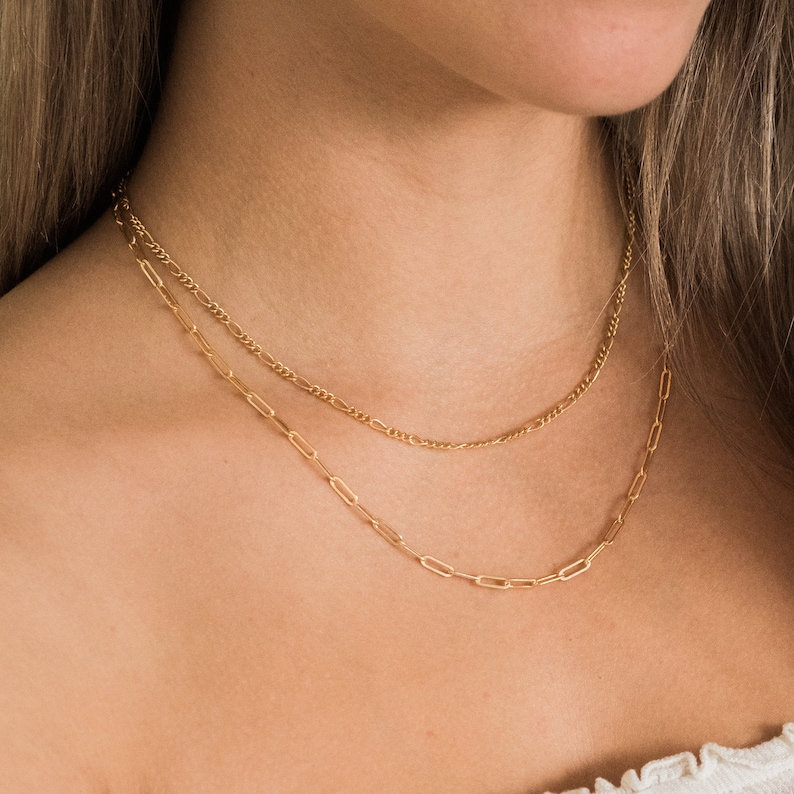 May include: Two gold necklaces layered on a person's neck. The top necklace is a delicate chain with small, evenly spaced links. The bottom necklace features a paperclip chain design with elongated rectangular links. The necklaces are a warm gold color.