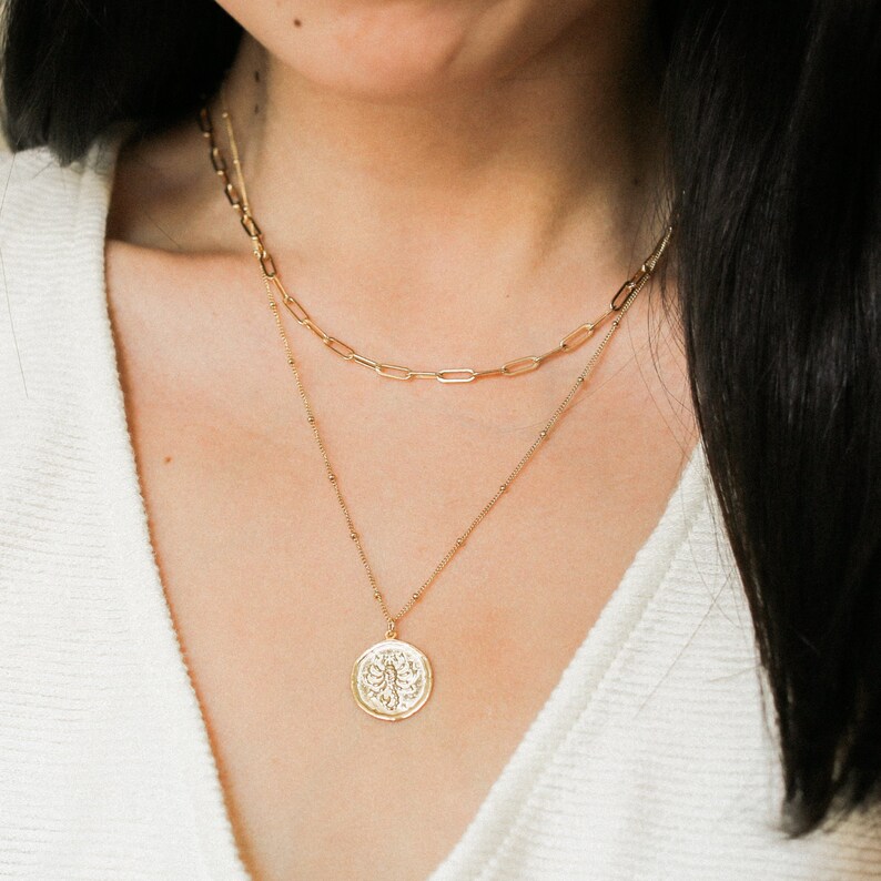 May include: A layered gold necklace set. The top layer is a chain link style, and the bottom layer features a pendant with a detailed design. The necklaces are worn over a white top.