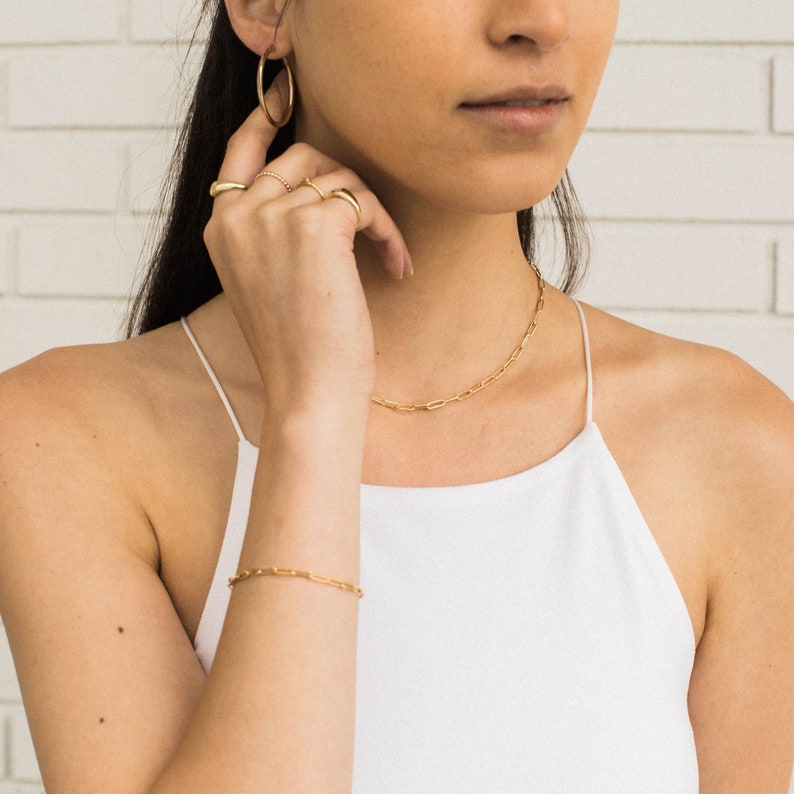 May include: A collection of gold jewelry, including a necklace, bracelet, hoop earrings, and rings. The jewelry is displayed on a person wearing a white tank top. The background is a white brick wall.