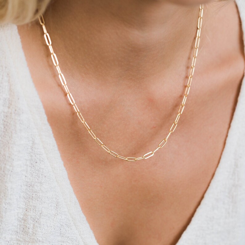 May include: A delicate gold-colored necklace with a paperclip chain design. The necklace is made of small, rectangular links. The jewelry is worn around the neck and rests on a white garment.