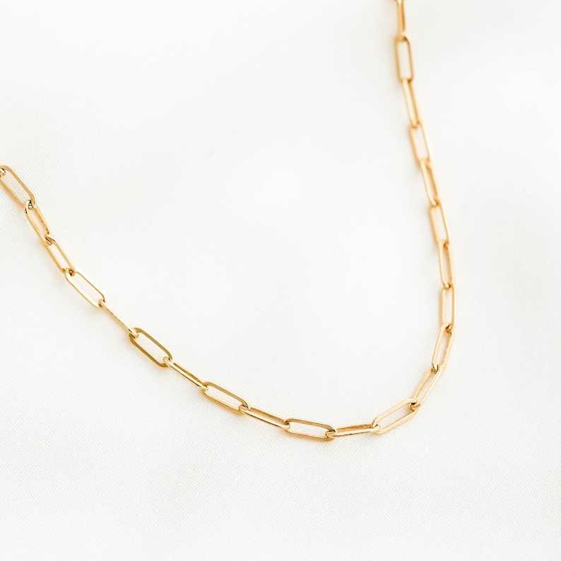 May include: A gold-colored paperclip chain necklace. The necklace features a series of elongated, rectangular links. The chain is displayed against a white background, highlighting its simple, elegant design. This jewelry piece is suitable for everyday wear.